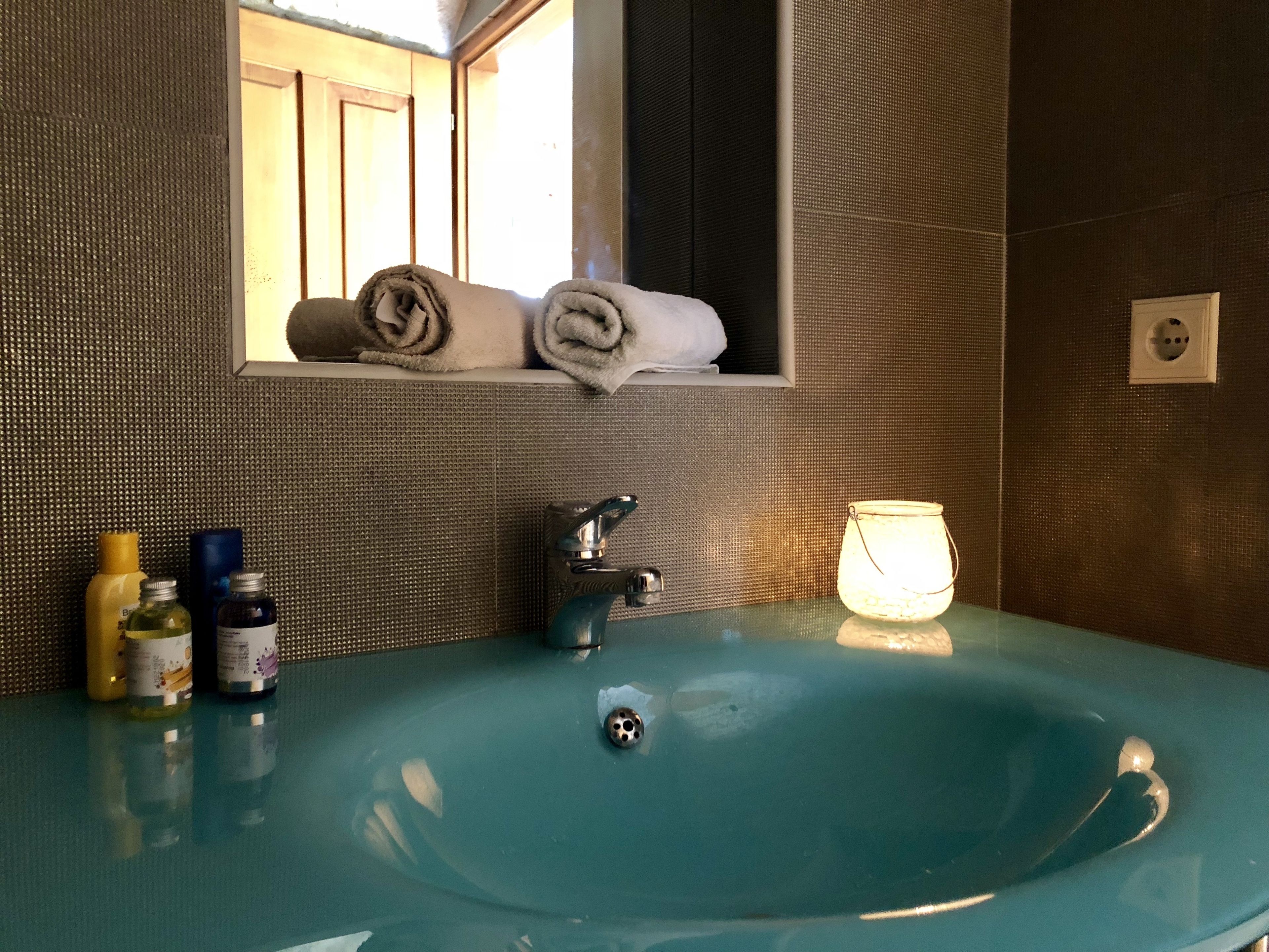 A bathroom with a teal sink, a mirror, and a window. There are towels stacked in the mirror, a soap dispenser, and a glass jar on the sink.