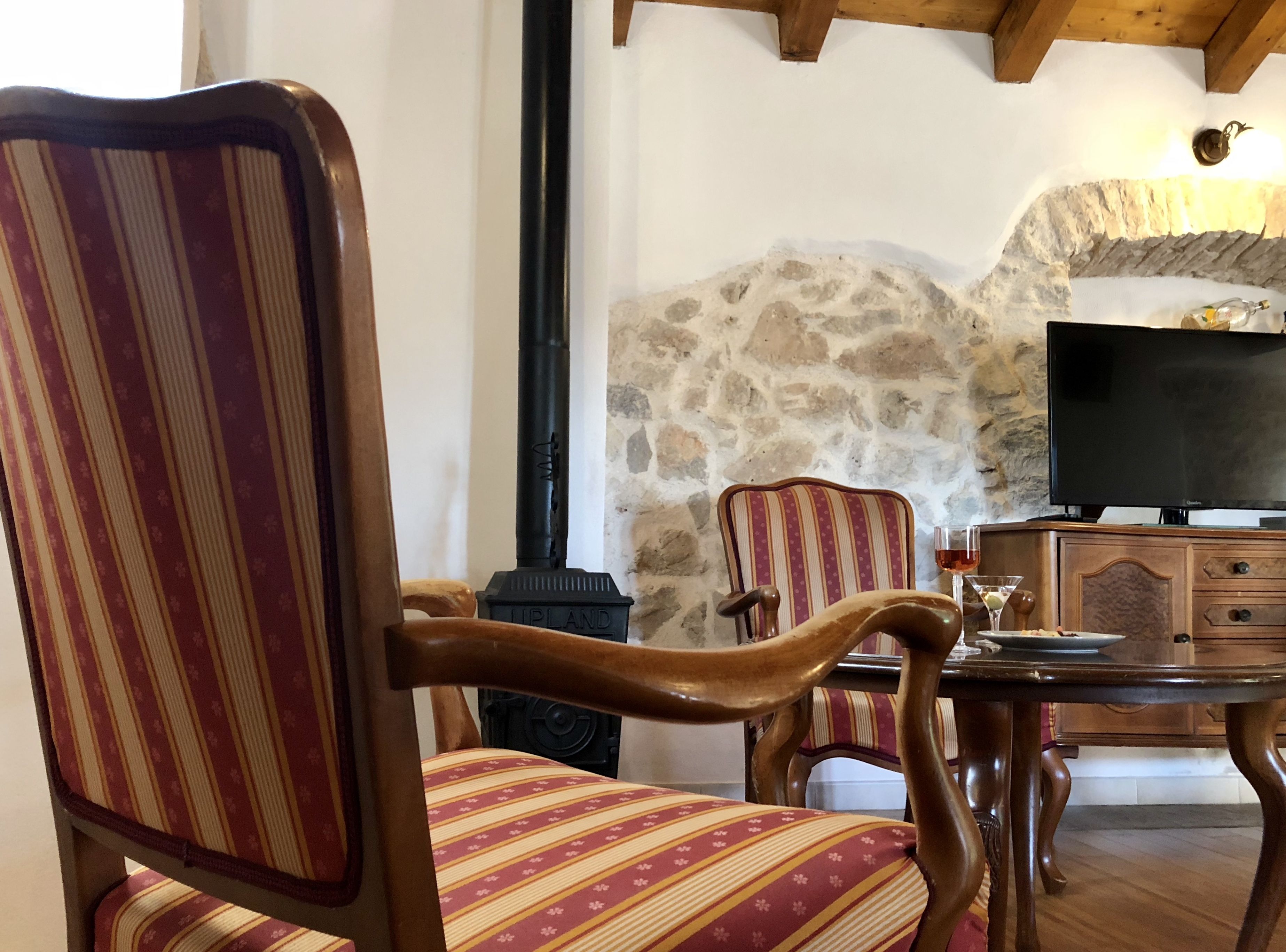 A cozy living room with striped upholstered chairs, a stone wall, a fireplace, and a television.
