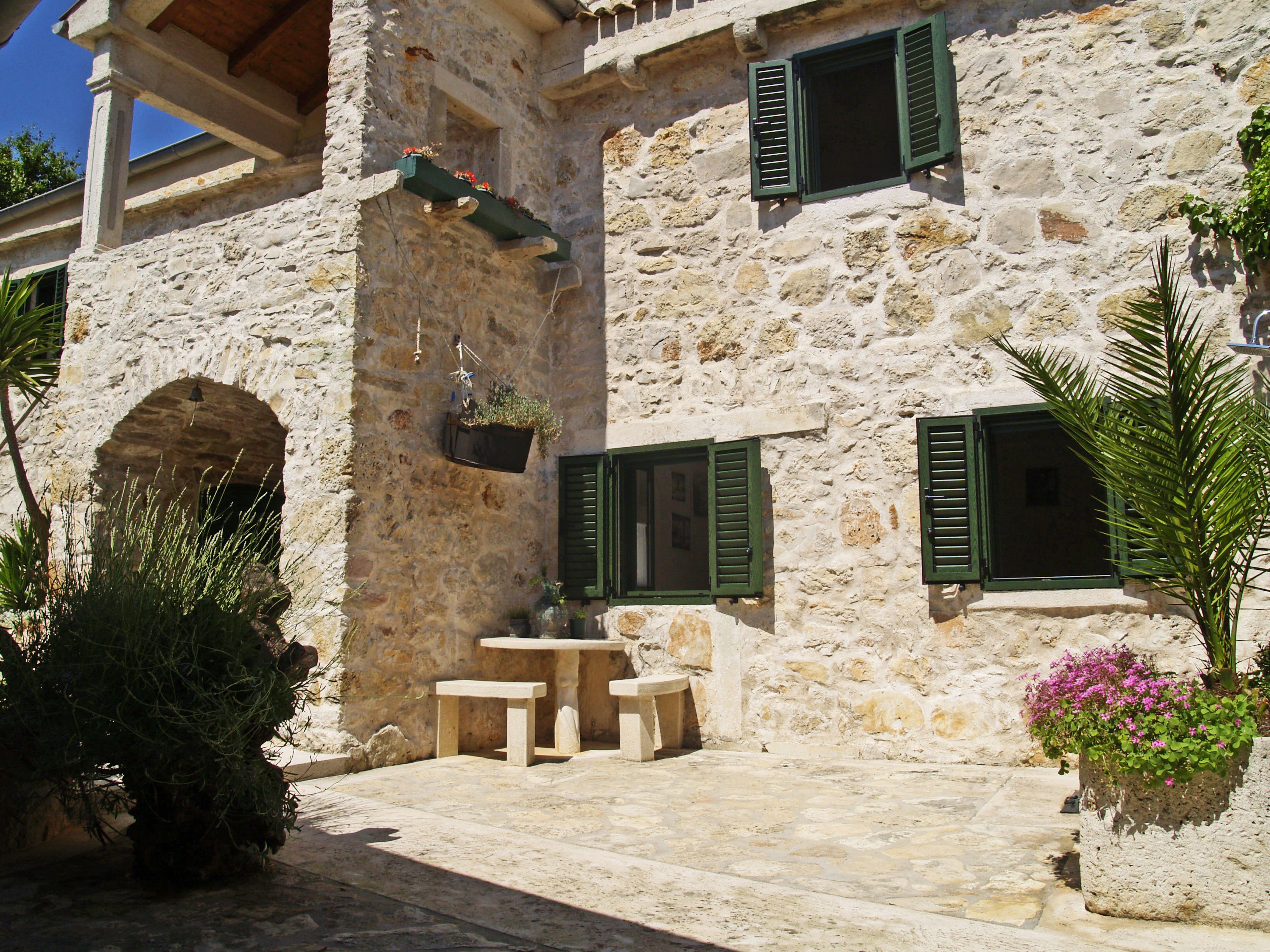 A stone building with green shutters, a small outdoor seating area, and potted plants.