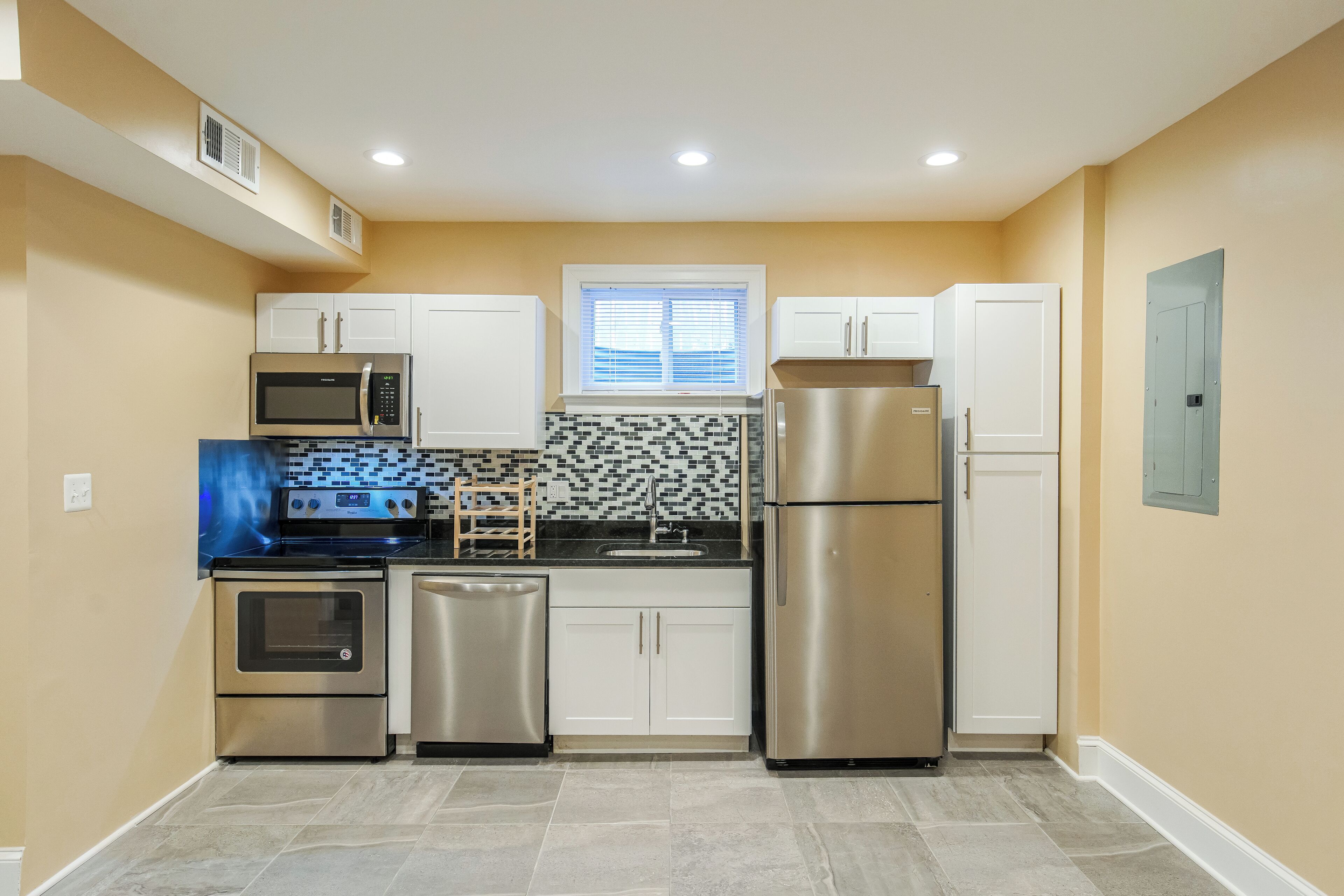 A compact kitchen with white cabinets, stainless steel appliances, a patterned backsplash, and a window with blinds.