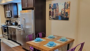 A compact kitchen with modern appliances, a dining table with purple chairs, and a cityscape painting on the wall.