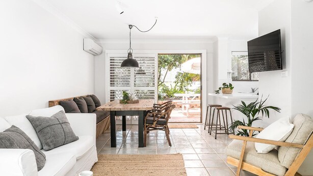 A modern living space with a white sofa, a dining table with chairs, a kitchen area with a TV, and a view of the outdoors. - A PERFECT STAY - Cooinda  (Byron Bay)