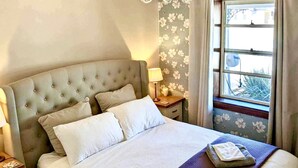 A neatly made bed with a tufted headboard, a bedside lamp, and a window with floral curtains. - Refurbished miners cottage 5 min from Moonta Bay + free internet (Moonta)