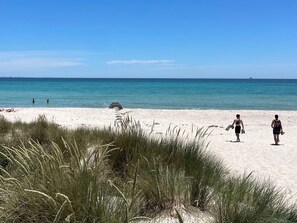 Ein Strand mit Sand und Meer, einige Personen und hohes Gras im Vordergrund.