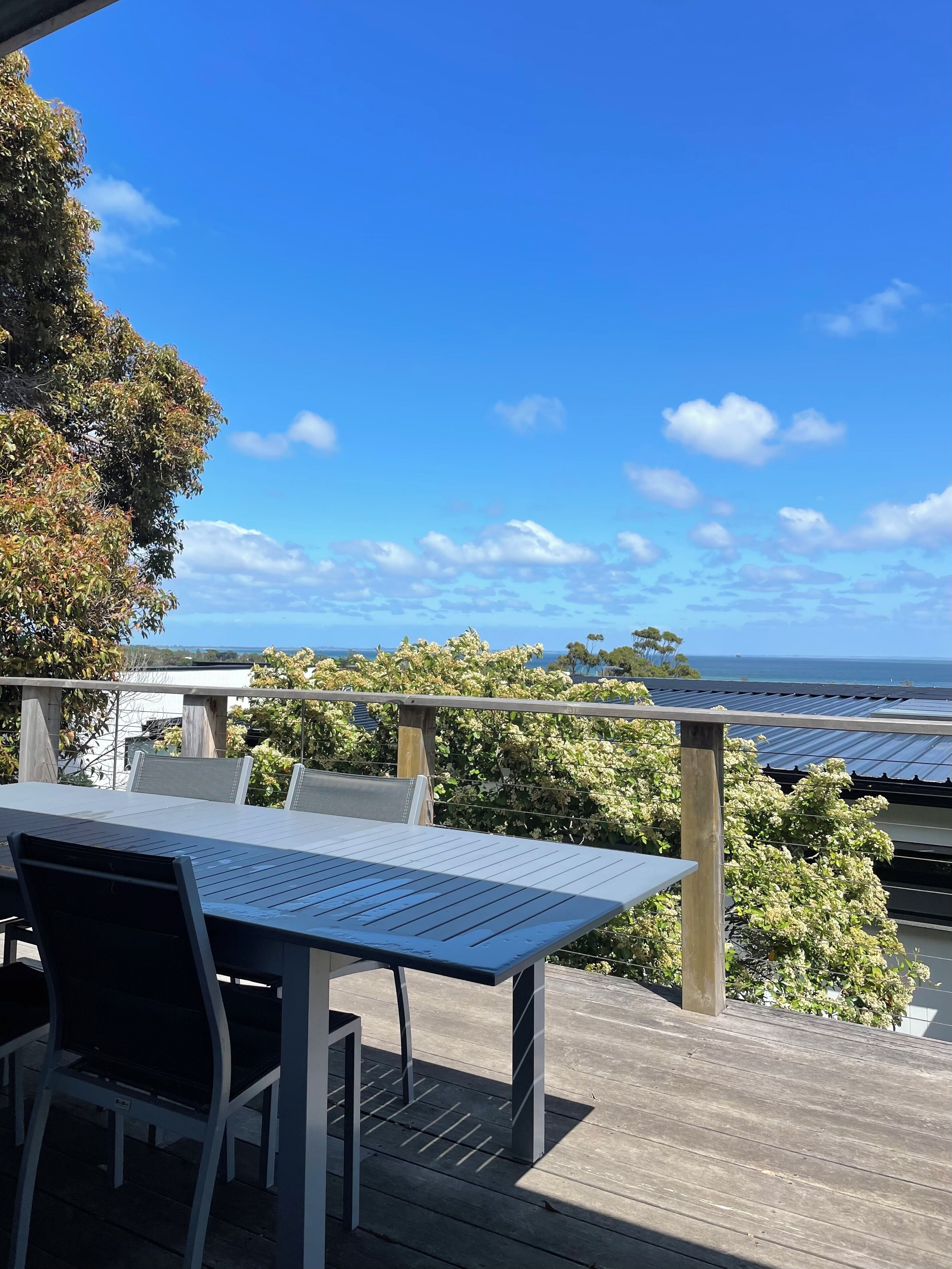 A wooden deck with a table and chairs, overlooking a body of water and a clear sky.