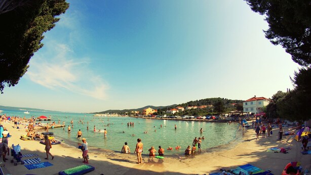 A crowded beach with people swimming and sunbathing, a clear sky, and buildings in the background. - Holiday Apartments Paloma (Pasman)