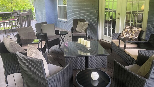 Een overdekt terras met rieten meubilair, een glazen tafel en een deur naar een groene tuin.