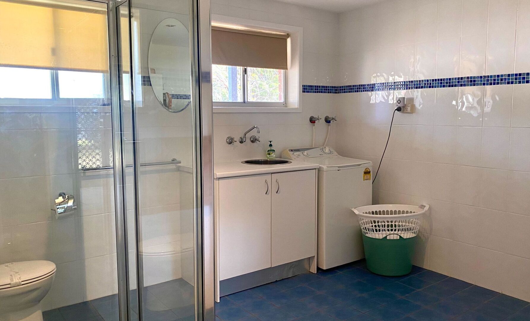 A bathroom with a glass shower enclosure, a toilet, a sink with a mirror, a washing machine, and a laundry basket.