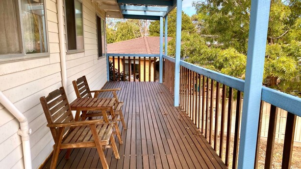 A wooden deck with two wooden chairs and a table, a blue railing, and a view of trees and a building in the background.