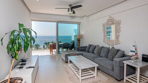 A modern living room with a grey sofa, a white coffee table, and a ceiling fan. A large window offers a view of the ocean. - Spectacular Beach front 2 Bedroom Condo (Riviera Nayarit)