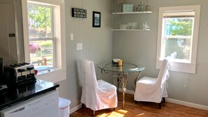 Una cocina con tostadora, refrigerador y comedor con dos sillas y una mesa de vidrio. Hay dos ventanas que dejan entrar luz natural, y un estante con objetos decorativos sobre la mesa.