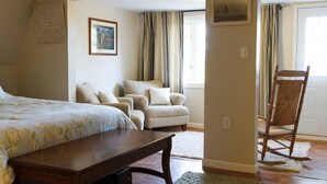 Un dormitorio con una cama, un banco de madera, una silla y un sillón mecedora. Hay dos ventanas con cortinas, un cuadro en la pared y un tapiz con textura.