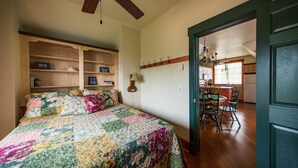 Una camera da letto con una coperta trapuntata, mensole di legno, una ventola a soffitto e vista sulla cucina tramite una porta aperta.