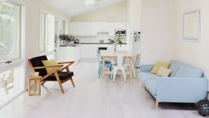Uma sala ampla com um sofá azul claro, uma cadeira de madeira, uma mesa de jantar com cadeiras e uma cozinha ao fundo.