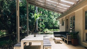 Uma área de varanda coberta com mesa e bancos de madeira, um sofá preto e um vaso com planta.