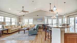 Een ruime woonkamer met een grote bank, een salontafel en een eethoek met stoelen en een kroonluchter. Er zijn plafondventilatoren en grote ramen die voor veel daglicht zorgen.