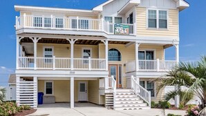 Een tweelaags strandhuis met een balkon en een overdekte veranda, met een bord waarop staat 'Smile n' Wave'.