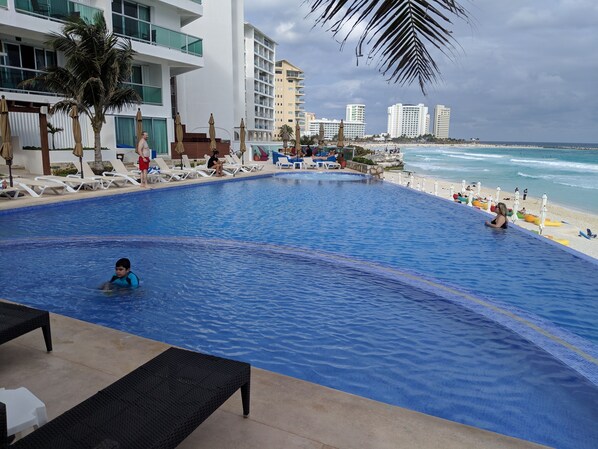 Una gran piscina al aire libre con sillones reclinables y sombrillas, rodeada de edificios modernos y una playa.