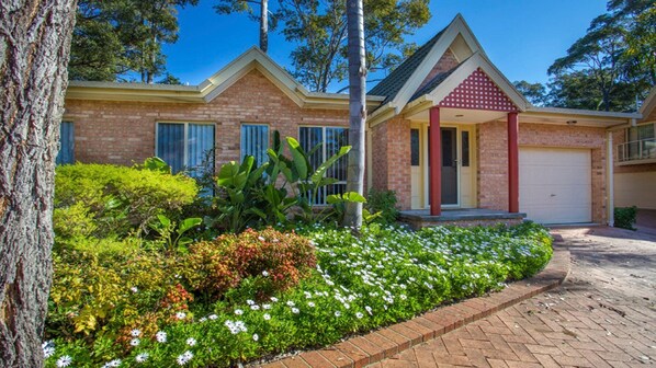 A two-story brick house with a garage, surrounded by a landscaped garden with various plants and flowers. - Unit Two in Denham Court- Stunning Beaches minutes away. (Denhams Beach)