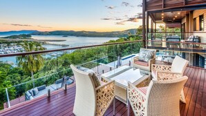 A balcony with wicker furniture and a glass table overlooking a scenic view of a lake and mountains during sunset.