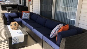 Un divano in vimini con cuscini blu e cuscini decorati, un tavolino intrecciato con una palla ornamentale, e un'area esterna riparata con vista sulla casa.