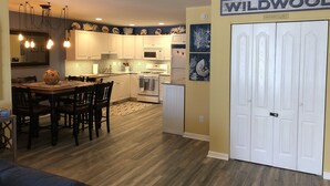 Una cucina moderna con mobili bianchi, una zona pranzo con tavolo e sedie in legno, e una porta chiusa con la scritta “WILDWOOD”.