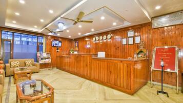 A hotel reception area with a wooden reception desk, a sofa, a coffee table, and a TV screen.