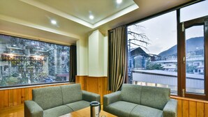 A room with two green upholstered chairs, a wooden table with a thermos and glasses, and large windows offering a view of a town and mountains. -  The Posh Hotel near Mall Road with Hassle Free Covered Parking (Dharamshala)