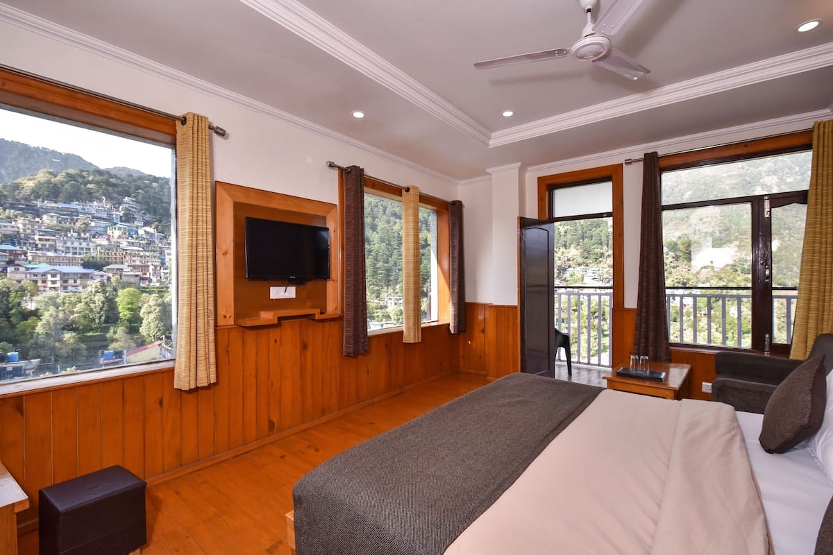 A hotel room with a large bed, wooden walls, a TV mounted on the wall, and a balcony with a view of the city.