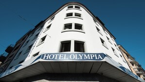 A corner view of Hotel Olympia with a prominent sign, multiple windows, and a clear blue sky. - Hotel Olympia (Zürich)