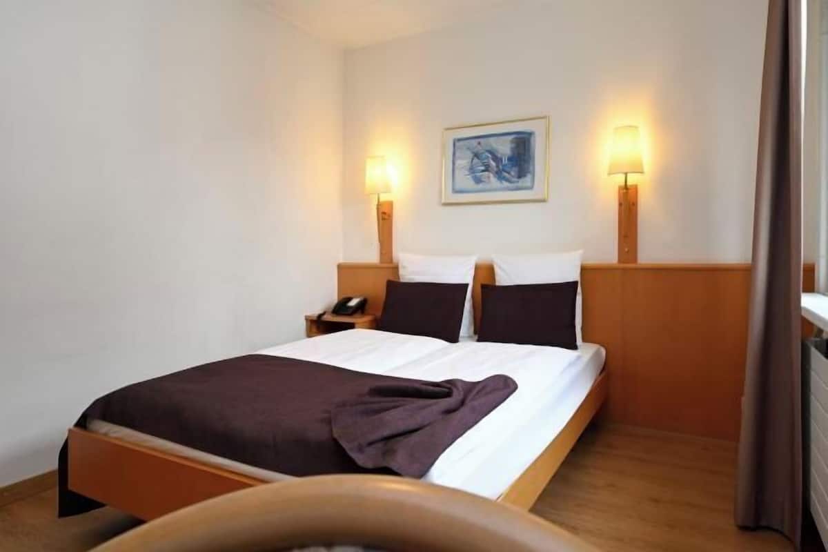 A hotel room with a bed, bedside tables, a lamp, a painting on the wall, and a window with curtains.