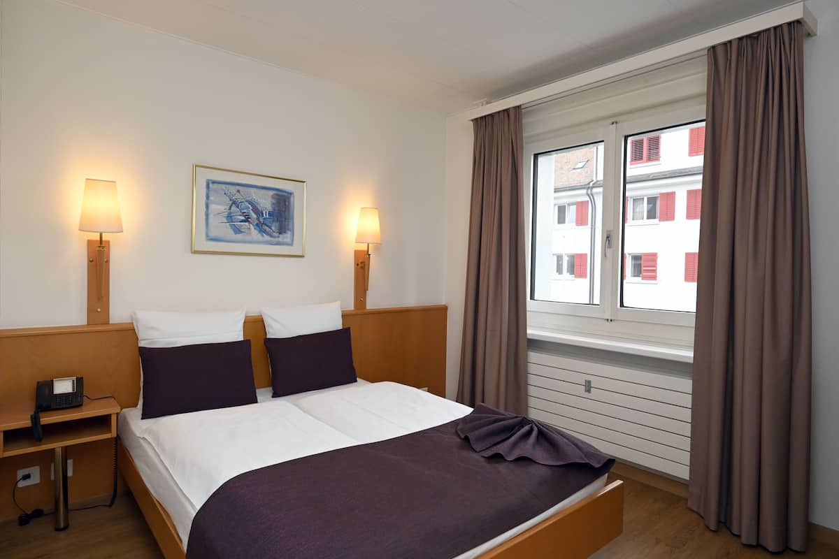 A hotel room with a bed, bedside lamps, a nightstand, a window with curtains, and a painting on the wall.