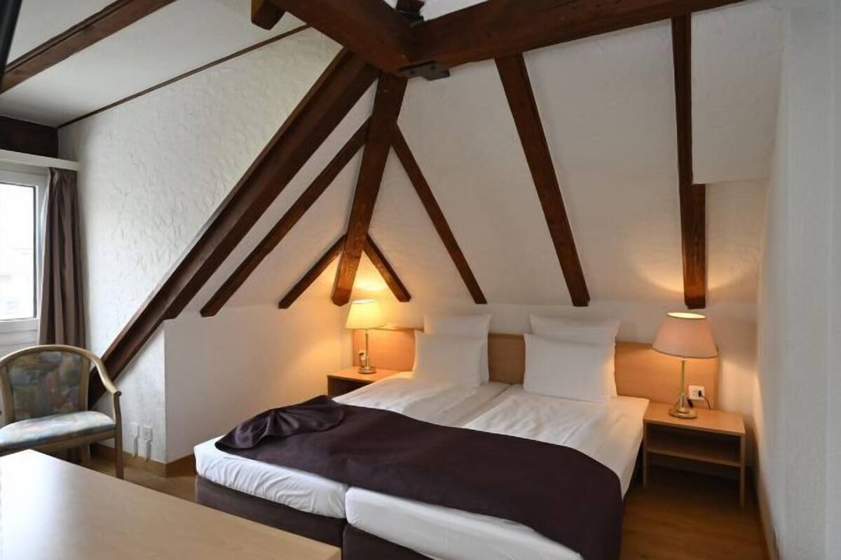 A bedroom with a sloped ceiling, a bed, two bedside tables with lamps, a chair, and a window with curtains.