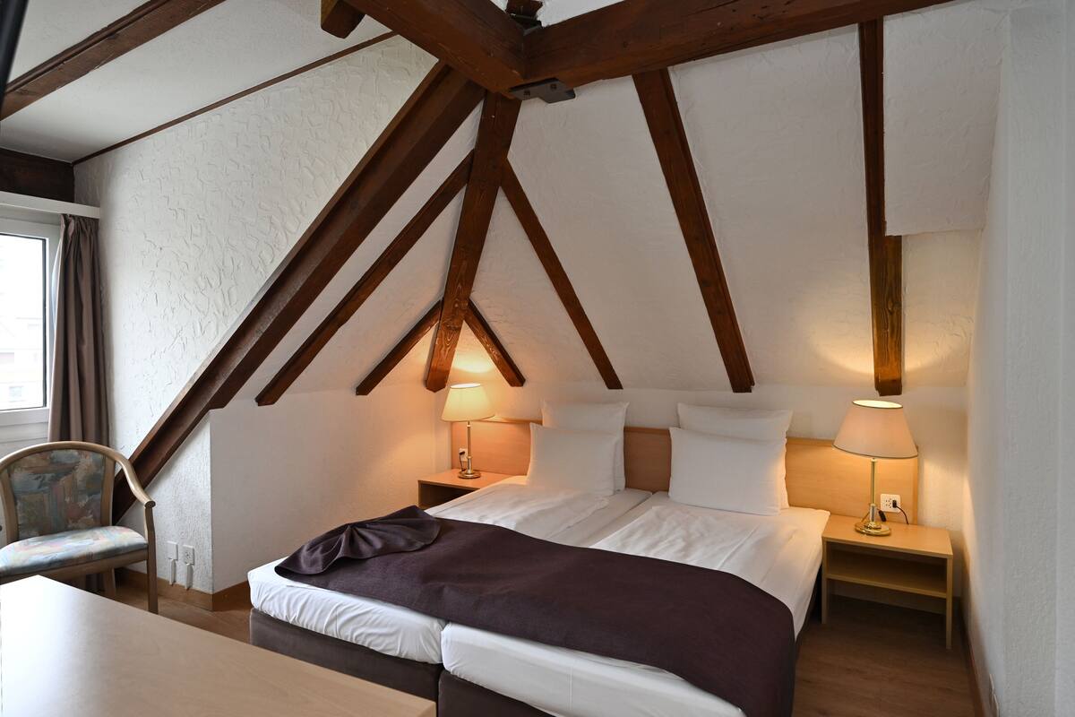 A bedroom with a sloped ceiling, wooden beams, a bed with white linens, two bedside tables with lamps, and a chair.