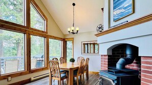 A dining area with a wooden table and chairs, large windows, a fireplace, and a ceiling light fixture. - Tree Top Cabin with access to Flathead Lake (Rollins)