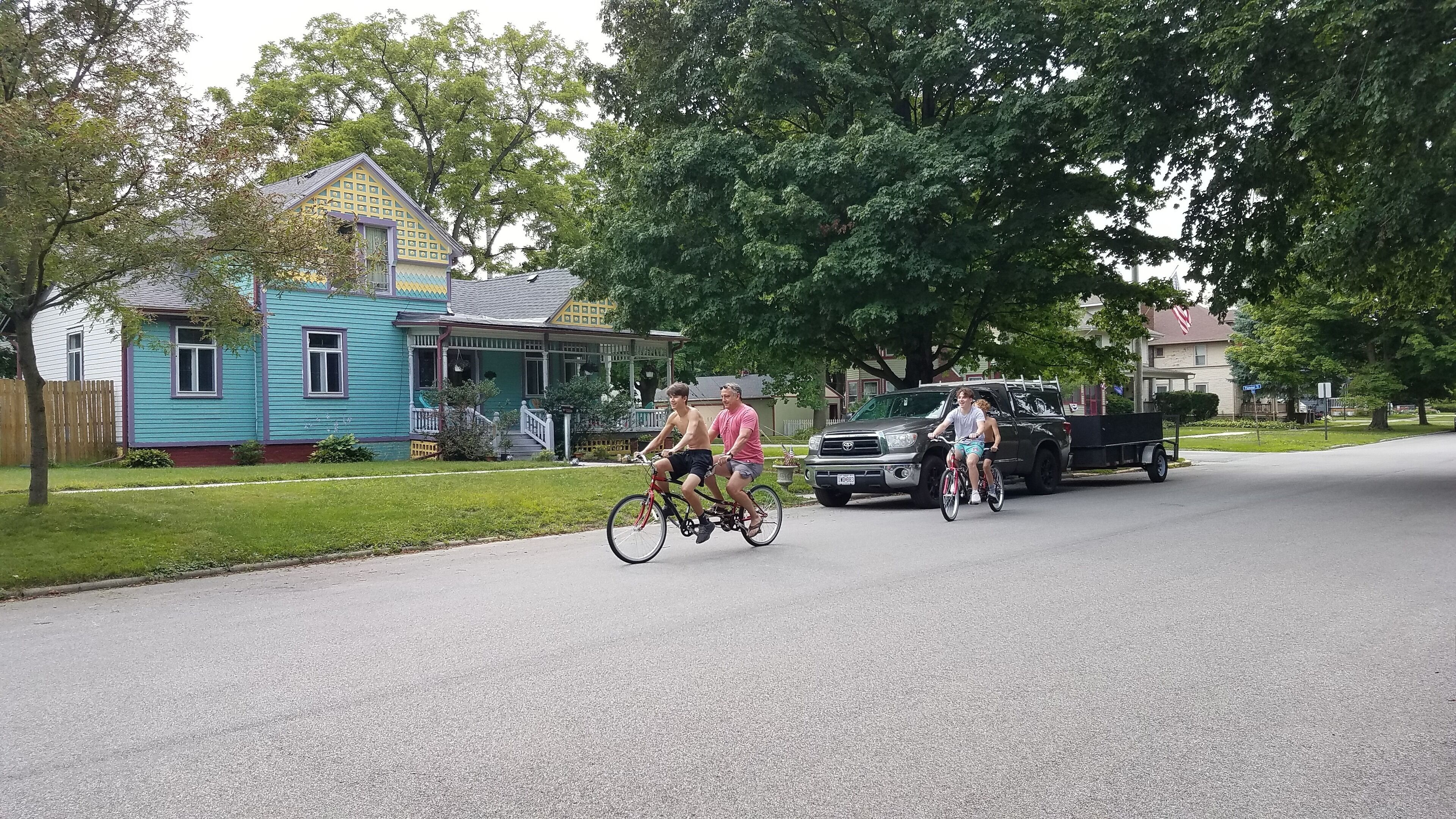 A residential street with houses, a parked truck, and cyclists.