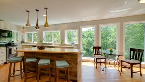 A modern kitchen with a central island, bar stools, a dining table with chairs, and large windows offering a view of the outdoors.