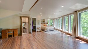 A spacious bedroom with a large bed, wooden flooring, and a view of greenery through large windows.