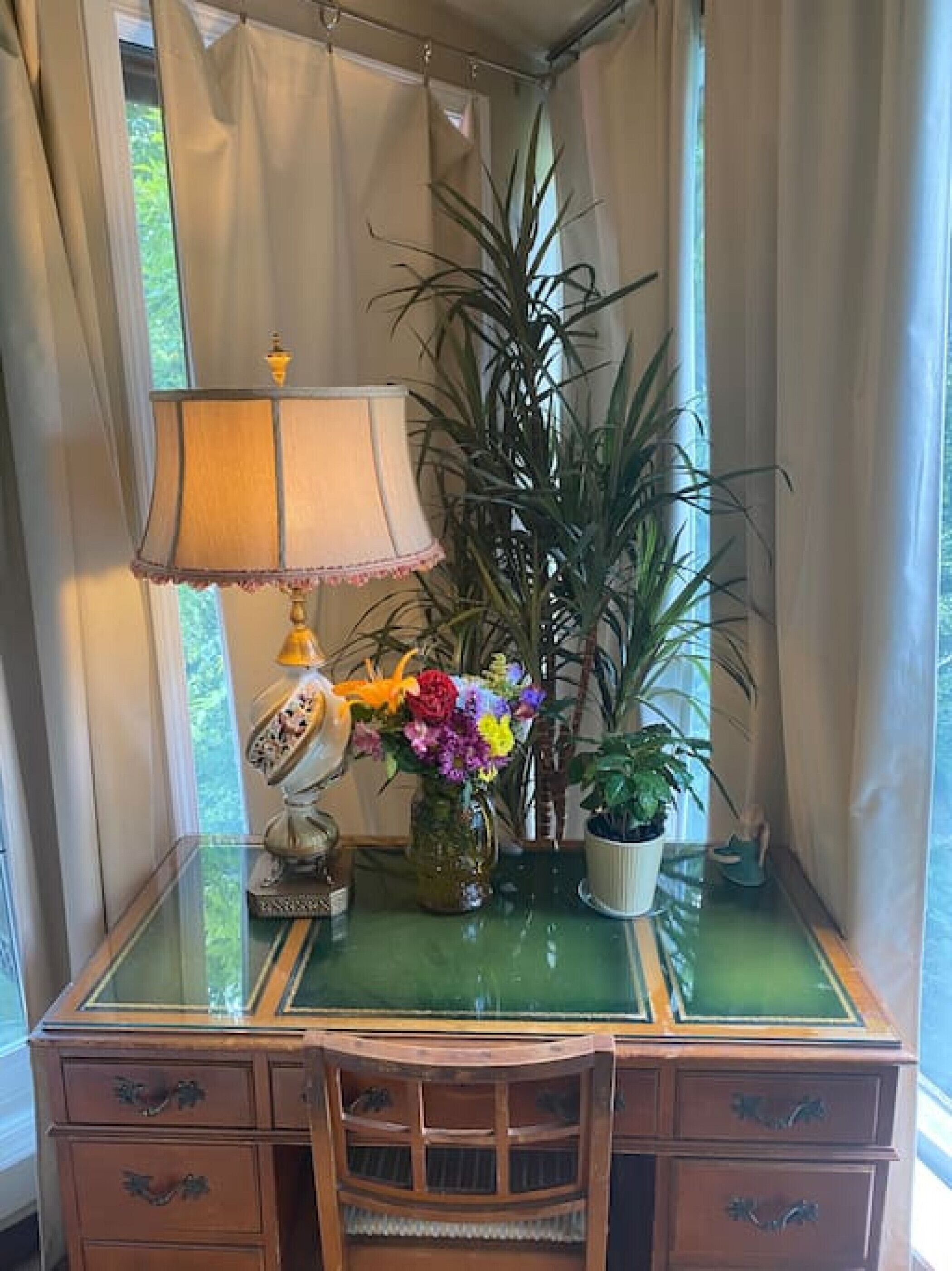 Un escritorio de madera con cubierta de vidrio, una lámpara, un florero con flores y una planta alta.