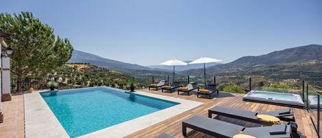 A pool area with lounge chairs and umbrellas, surrounded by a wooden deck and overlooking a mountainous landscape.