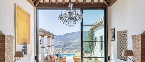 A spacious living room with a high ceiling, large windows, and a view of a pool and mountains.