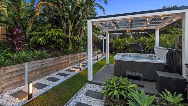 Uma área moderna com hidromassagem ao ar livre, com pérgola, cercada por plantas tropicais exuberantes e um caminho de cascalho.