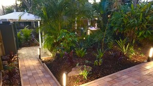 Um caminho no jardim com uma borda de tijolos, cercado por diversas plantas e um pequeno elemento aquático.