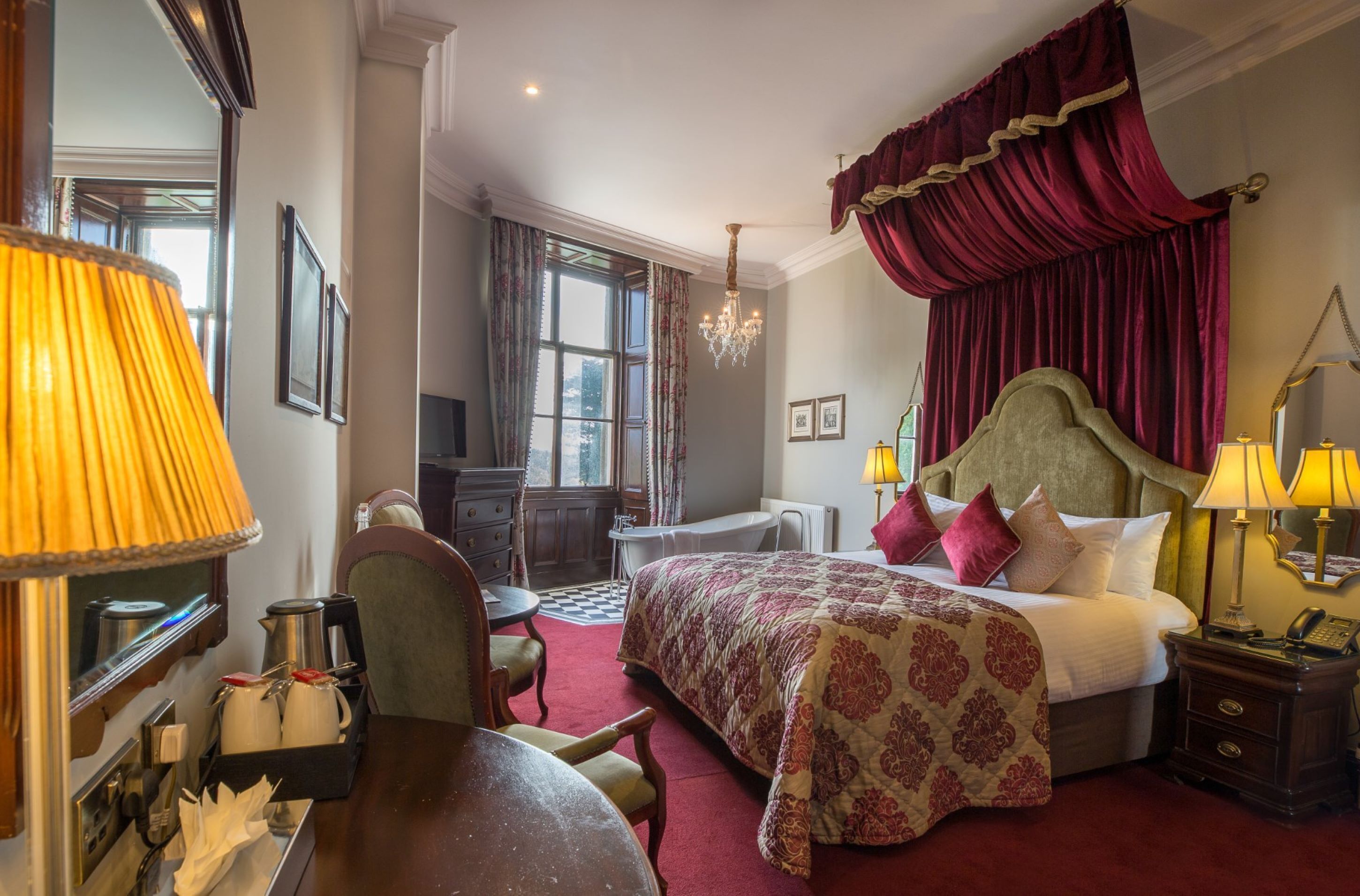 A hotel room with a large bed, a desk, a chair, a table, a chandelier, and a window with curtains.