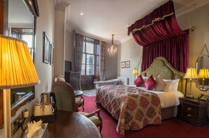 A hotel room with a large bed, a desk, a chair, a table, a chandelier, and a window with curtains.