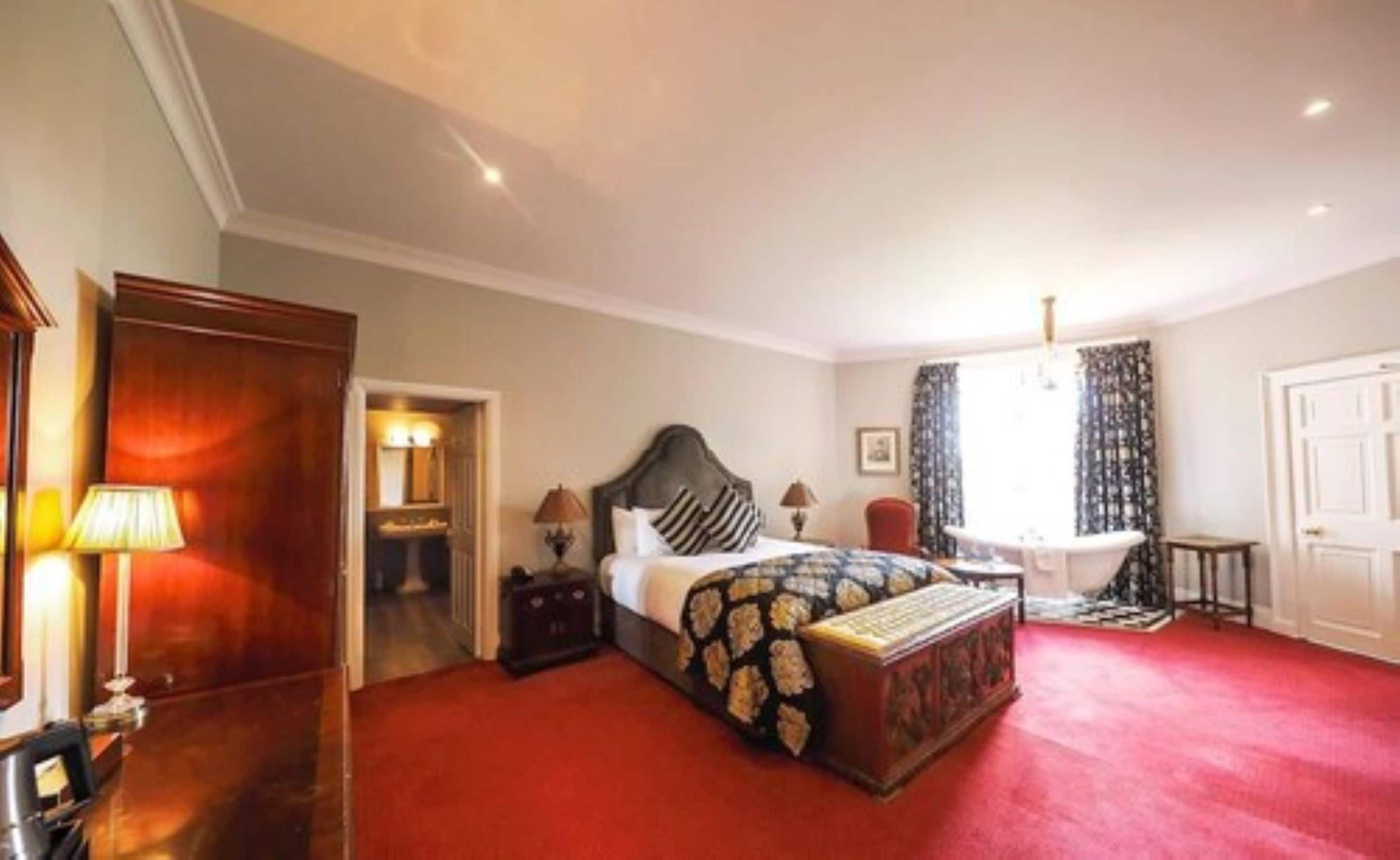 A hotel room with a large bed, a wooden headboard, a red carpet, a nightstand, a lamp, a dresser, a mirror, and a window with curtains.