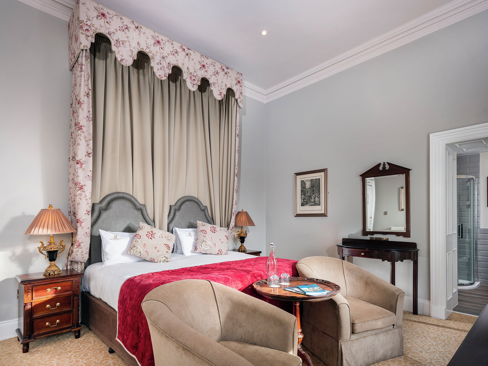 Een slaapkamer met een groot bed, twee nachtkastjes, een stoel, een klein tafeltje met dienblad, en een spiegel.
