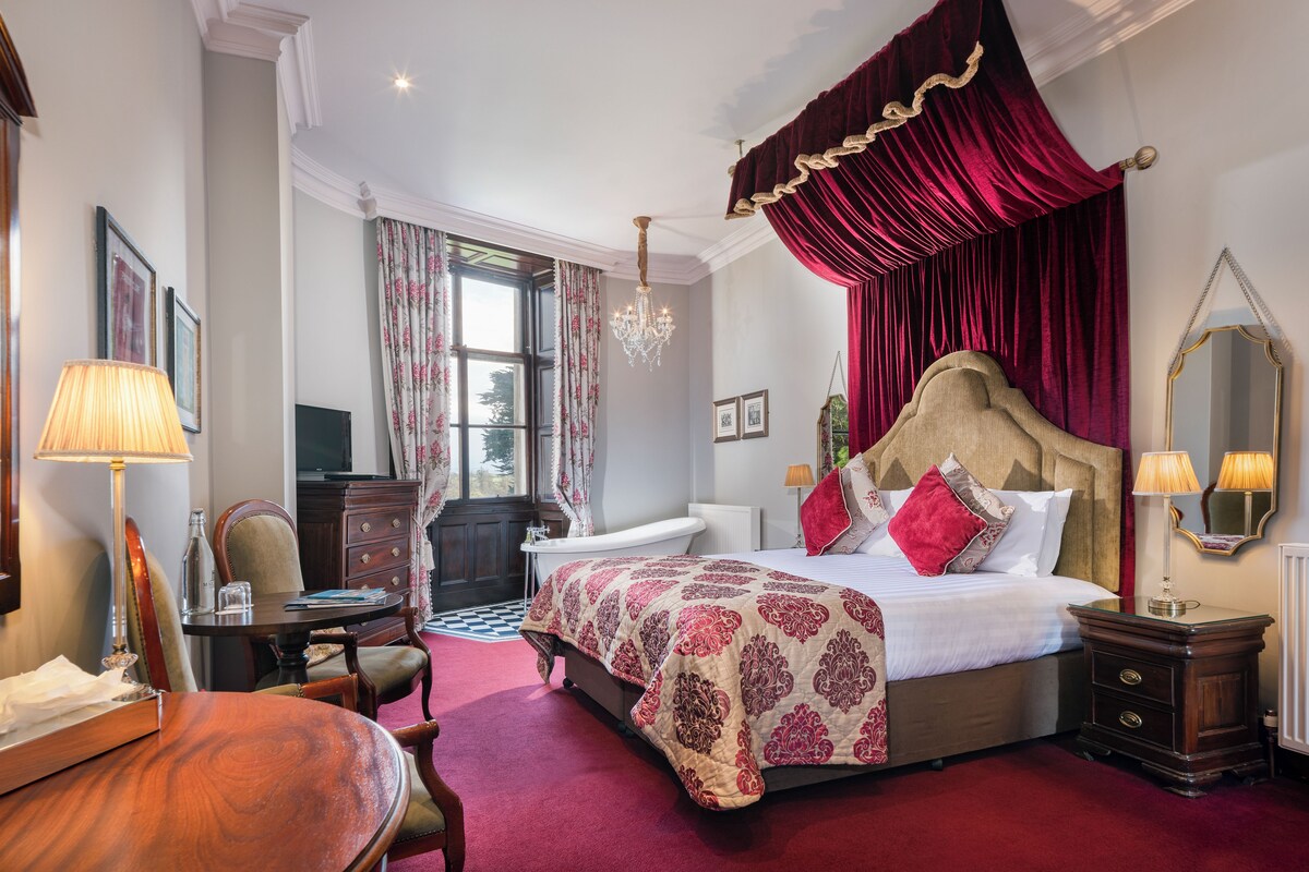 A bedroom with a four-poster bed, a desk with a chair, a chandelier, and a large window with curtains.