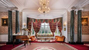 Une vaste pièce agrémentée d’un lustre, de colonnes richement décorées, d’un piano et d’une moquette rouge.