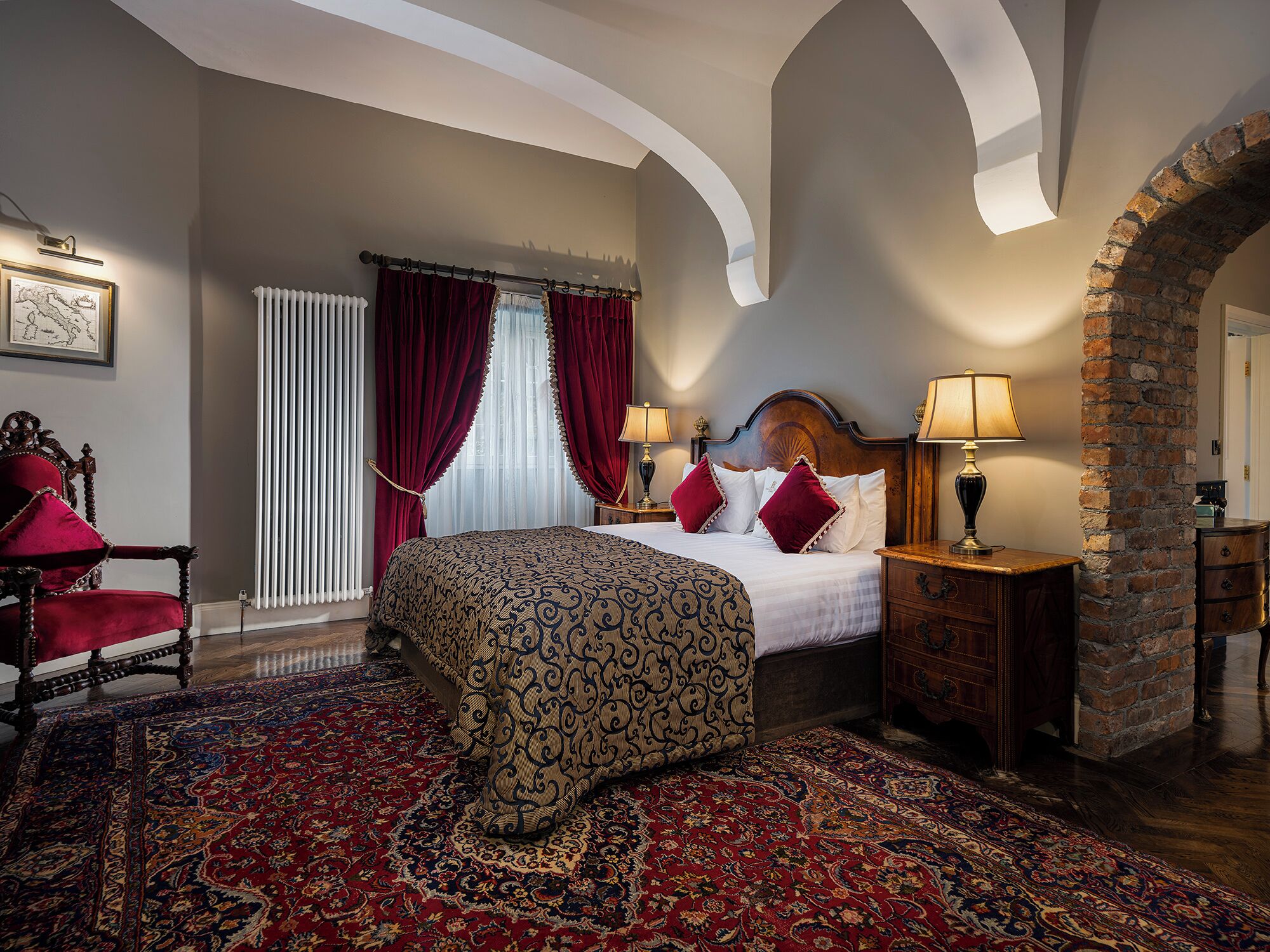 A hotel room with a large bed, a red and gold patterned carpet, a wooden chair, and a brick wall.